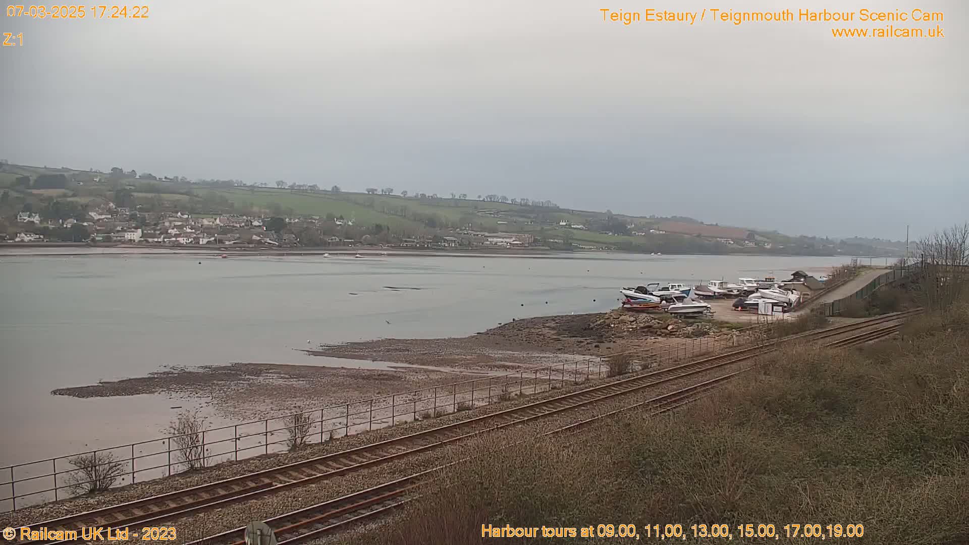 A wide shot shows a calm, overcast day overlooking a river with several boats docked near a train track.