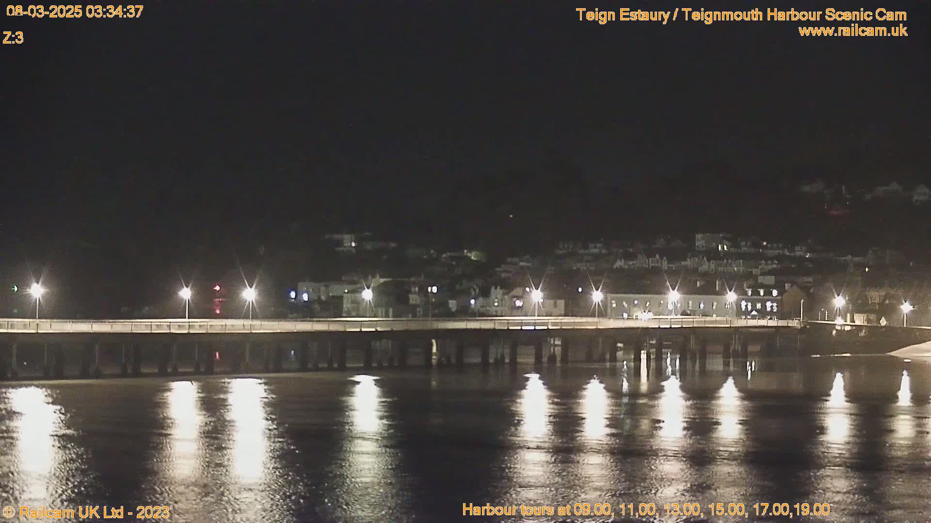 A long pier at night, illuminated by lights, stretches across calm, dark water reflecting the lights, with a town visible in the background under a clear, dark sky.