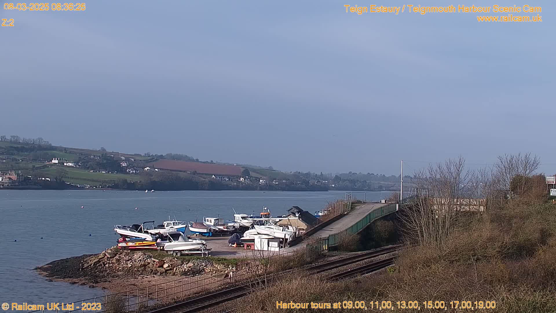 Several boats are docked at a harbor on a clear, sunny day, with a railway line and a bridge visible in the foreground.