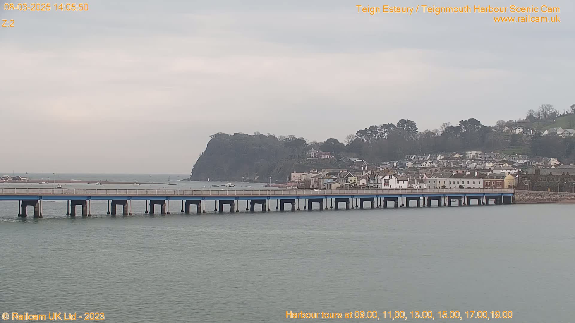 A long bridge spans a calm body of water under an overcast sky, leading to a town nestled against a hillside with a prominent cliff.