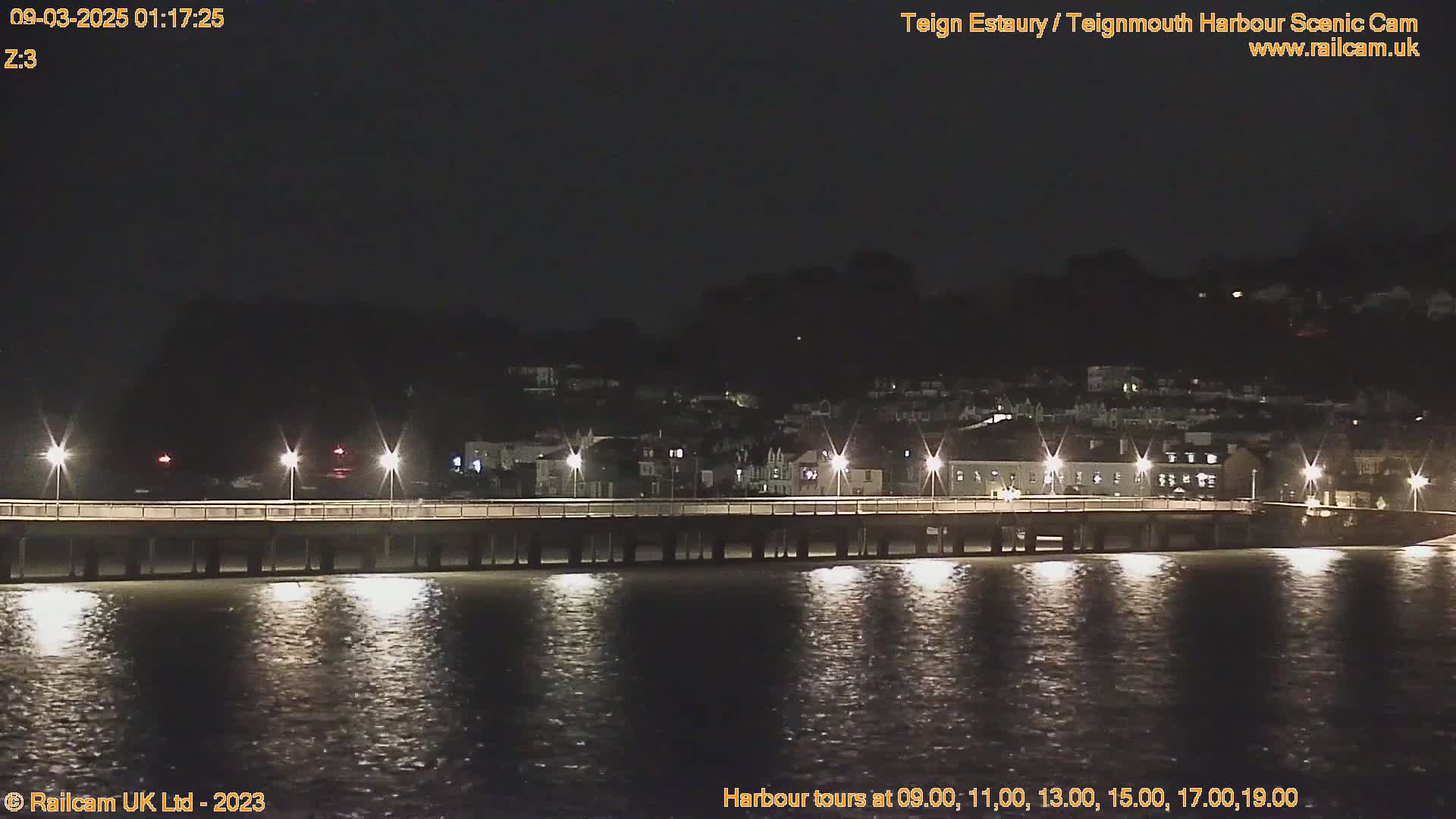 A long pier stretches across calm water at night, illuminated by lights, with a town rising on the far shore under a dark sky.