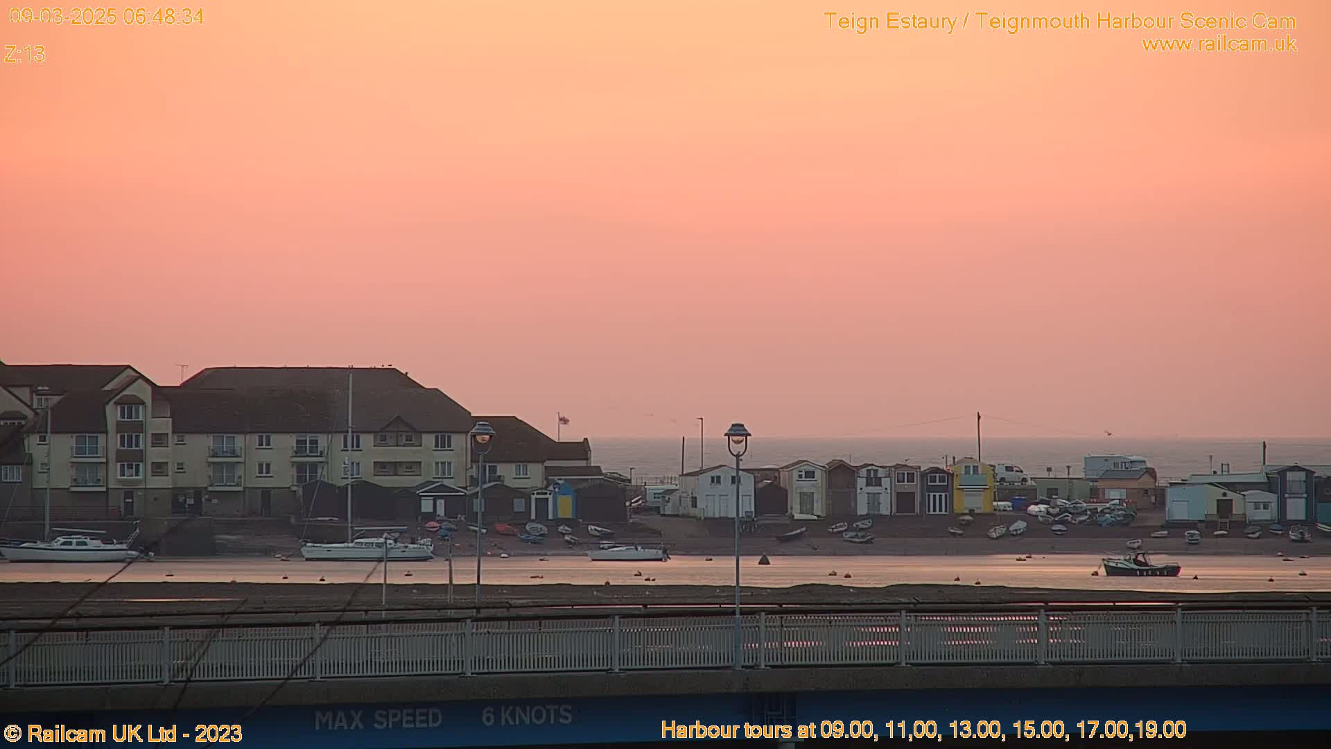 A pink sunrise illuminates a harbor scene with several boats and buildings along a calm body of water.