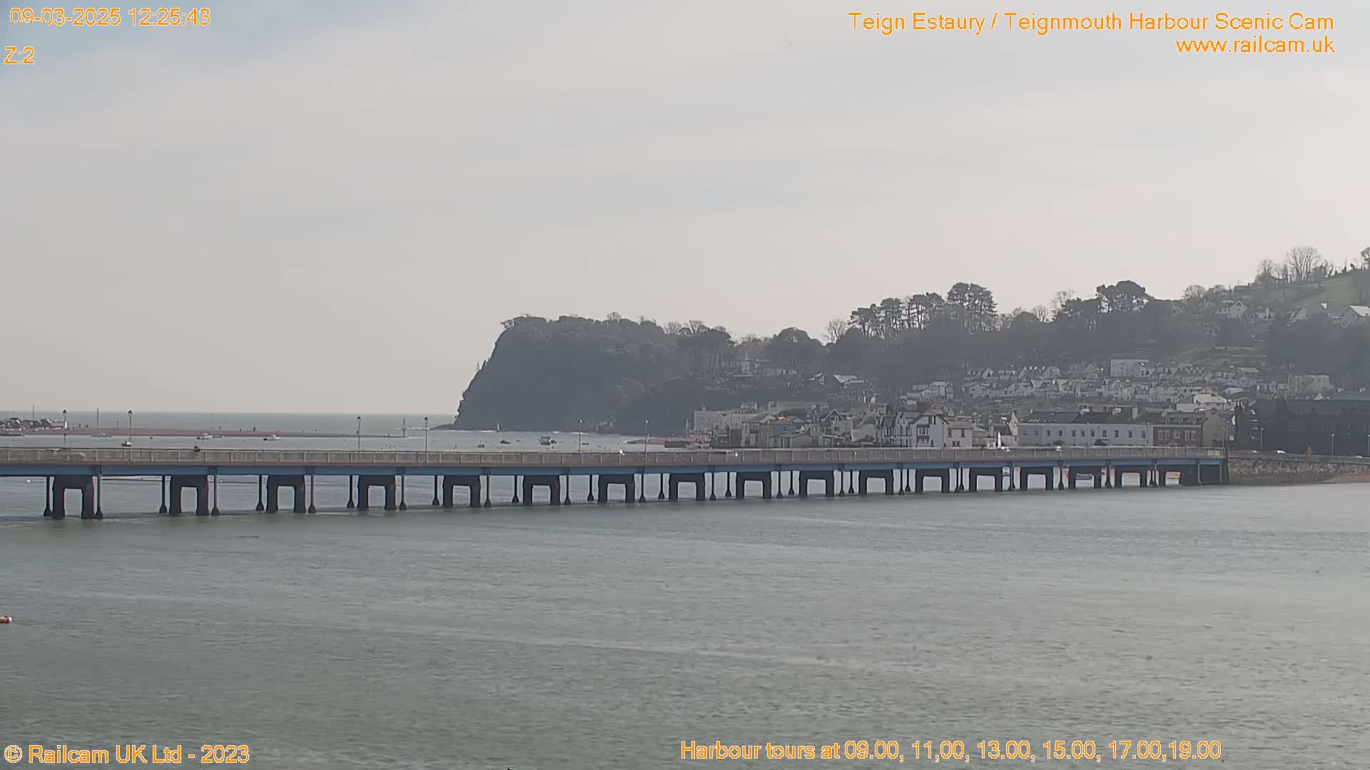 A long bridge extends across calm, gray water toward a town nestled against a hillside under an overcast sky.