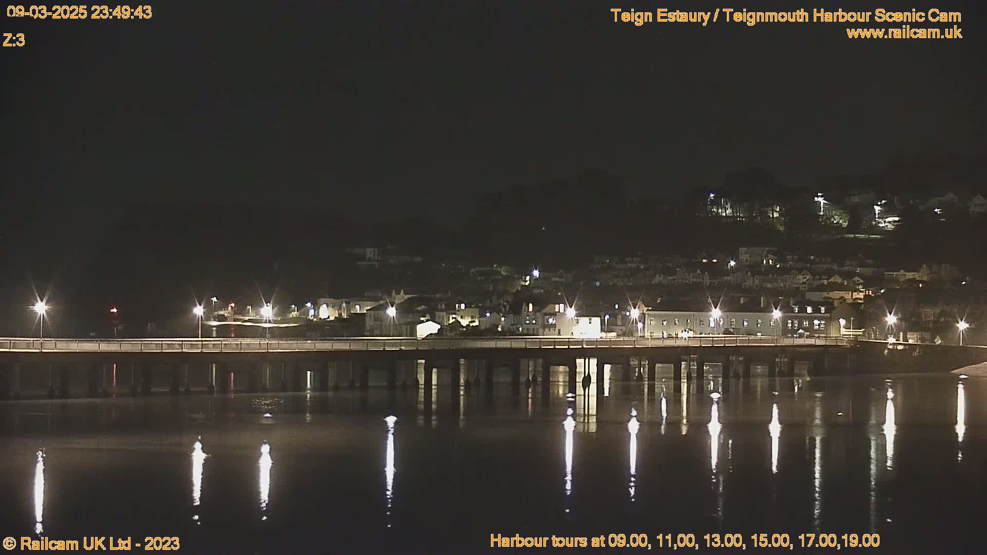 A nighttime view of a bridge over calm water, reflecting lights from buildings on the far shore under a clear, dark sky.