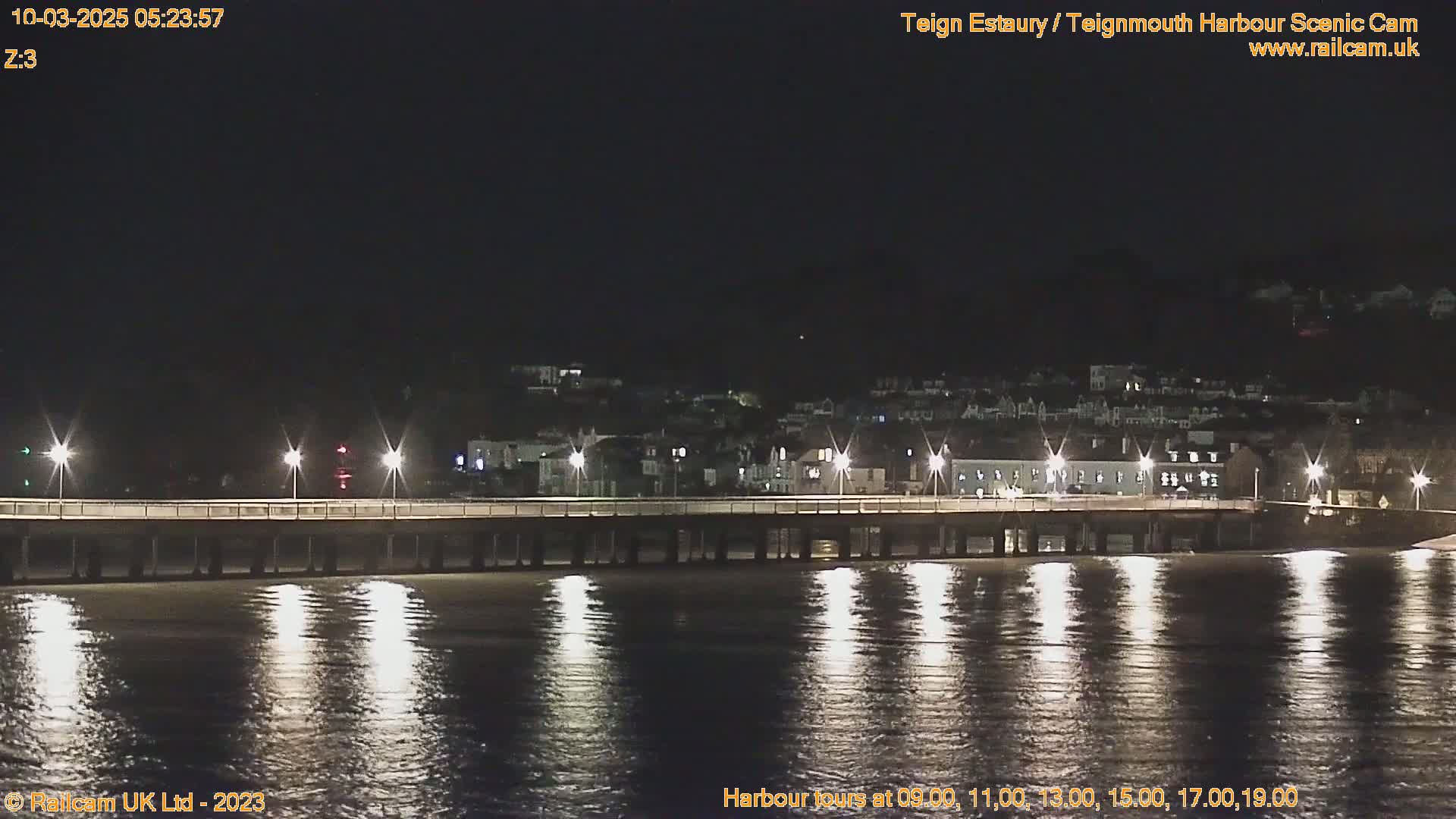 A long pier is illuminated at night, extending over calm, dark water reflecting the pier lights toward a town on the far shore.