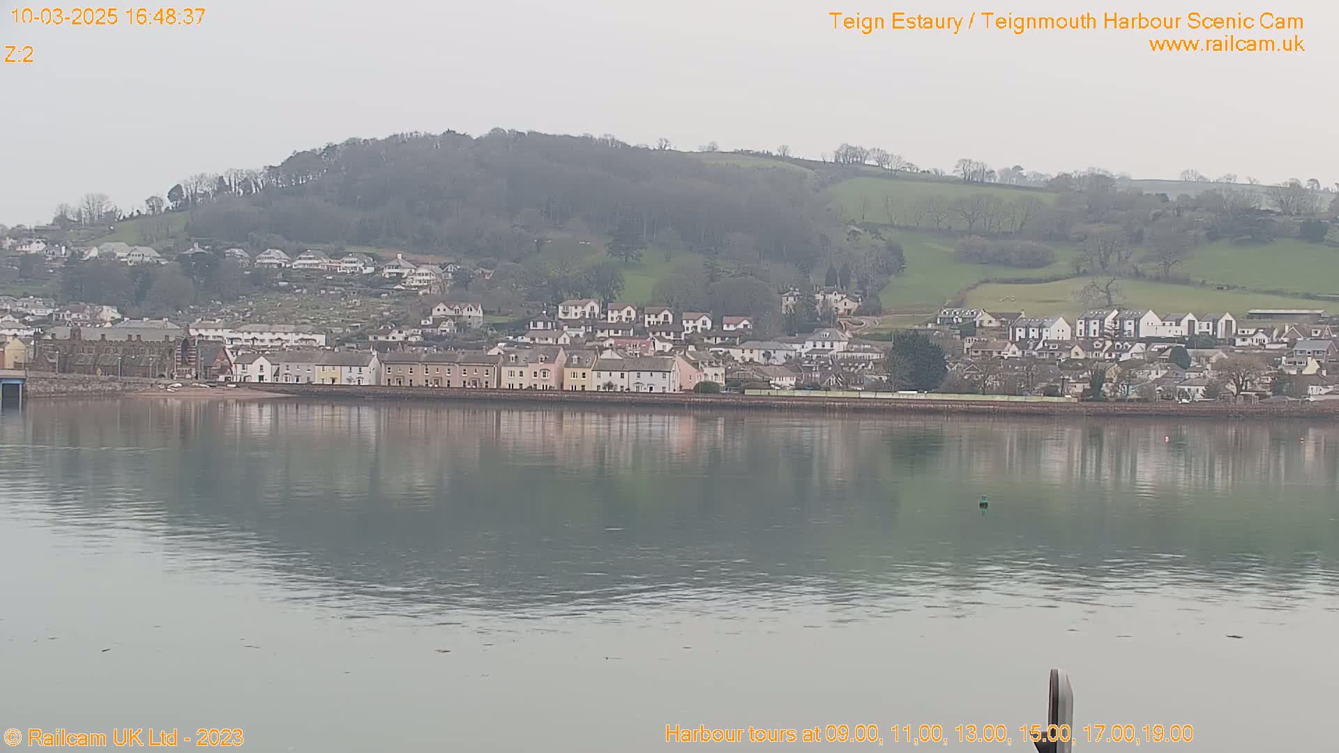 A calm body of water reflects a town nestled against a hillside under an overcast sky.