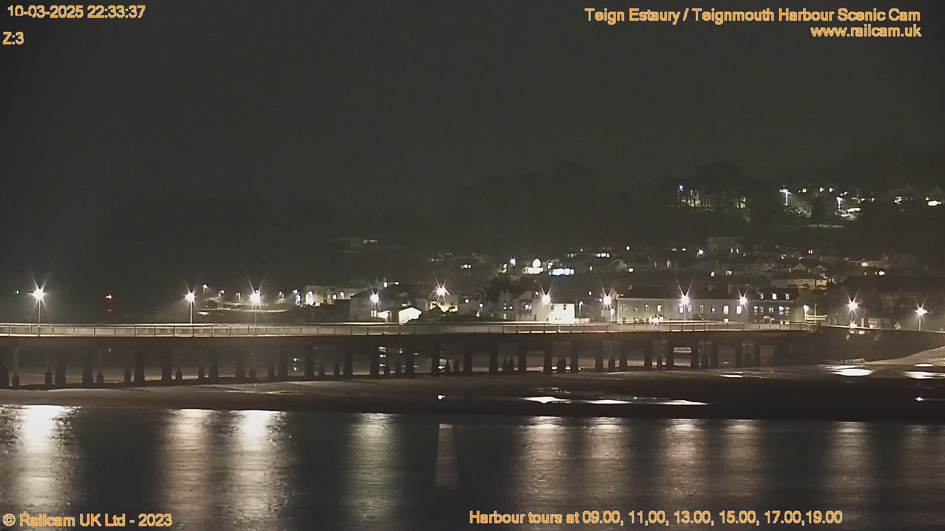 A long pier extends over calm, dark water at night, with a town's lights illuminating the far shore.