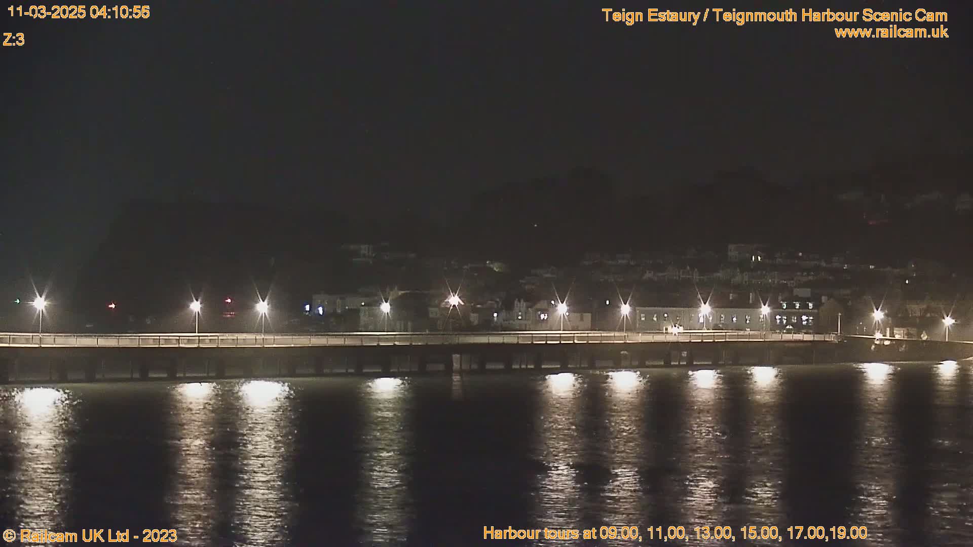 A long pier is illuminated at night, extending over calm, dark water towards a hilltop town.