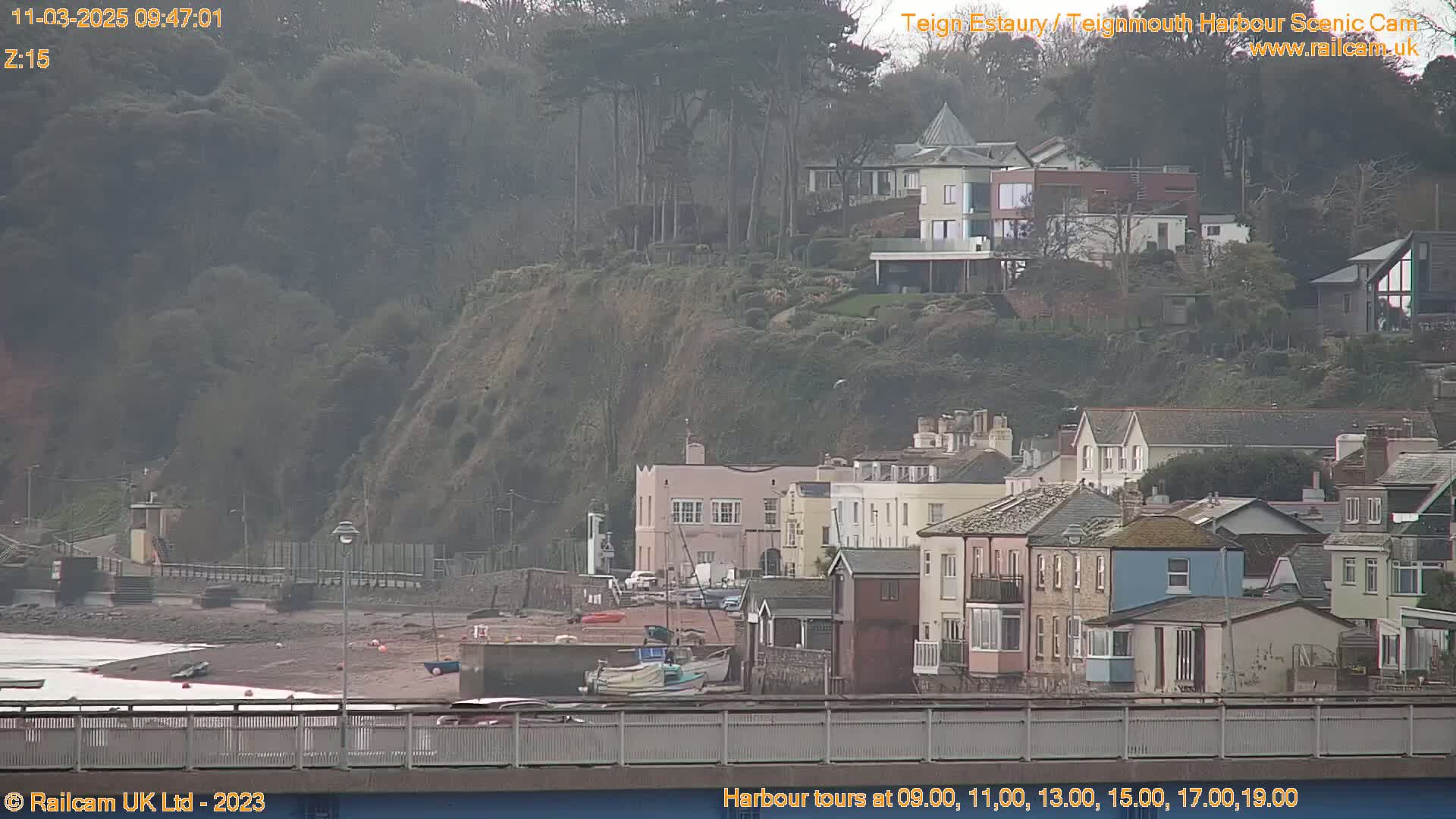A coastal town on an overcast day, with houses clustered near a rocky cliff overlooking a harbor where several small boats are moored.