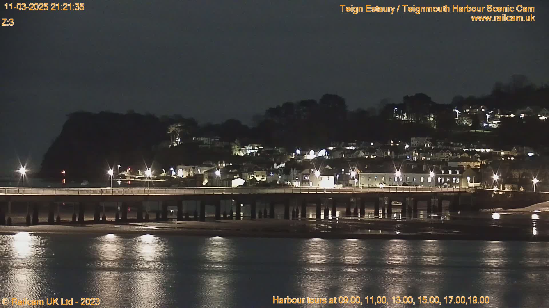 A nighttime view of a town situated on a hill overlooking a calm body of water with a long bridge extending from the shore, illuminated by streetlights.