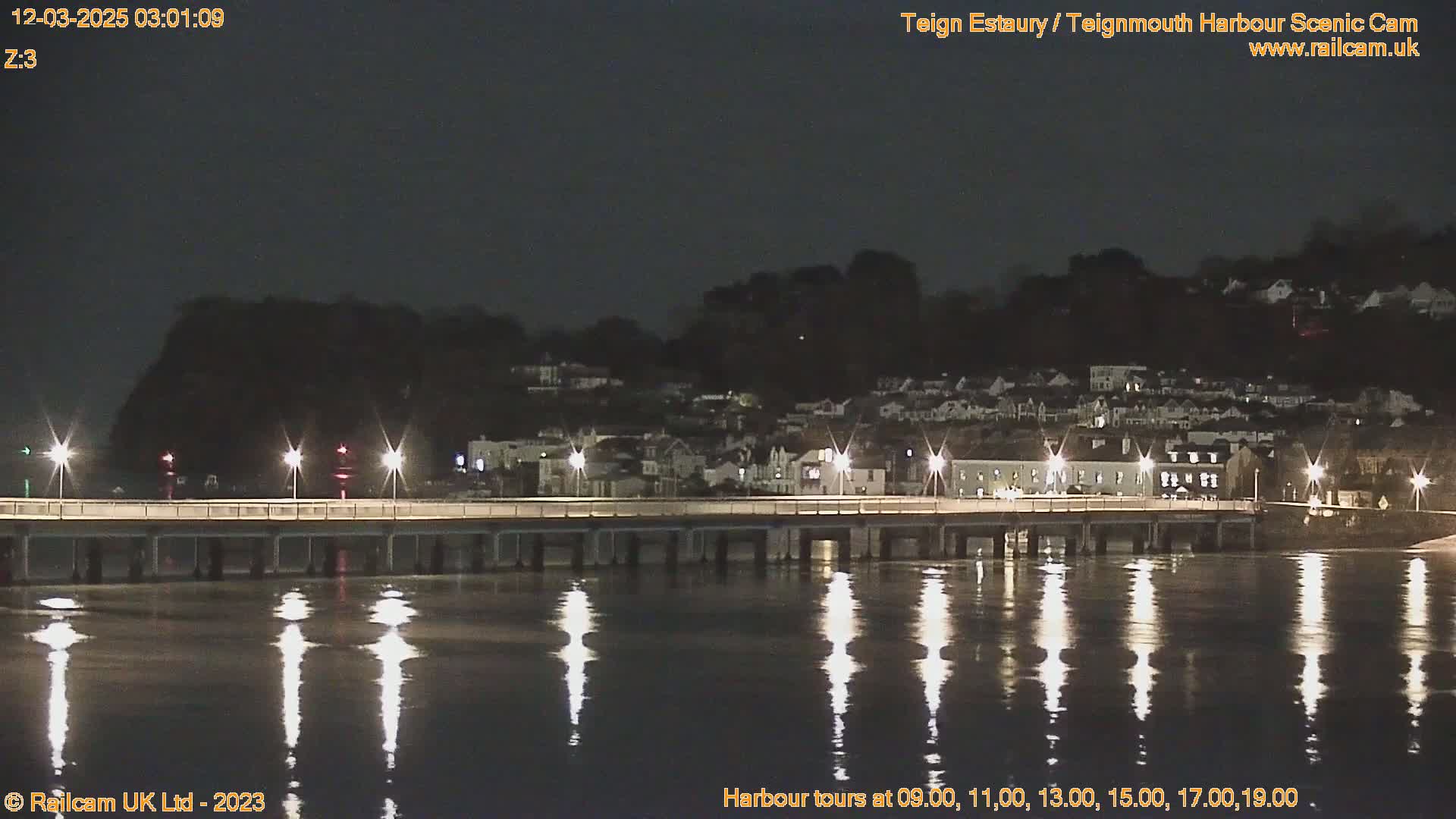 At night, a bridge illuminated by lights stretches across calm water reflecting the lights, with a hillside town visible in the background under a dark sky.
