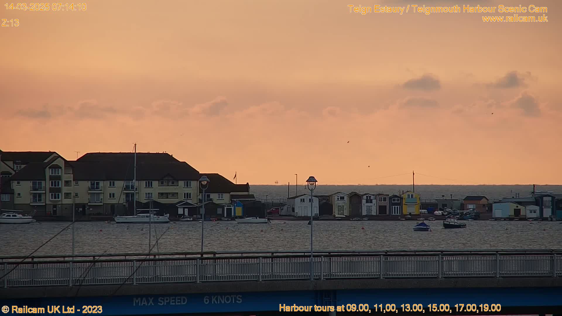 A calm harbor scene at sunrise, showing buildings along the waterfront and several small boats moored in the water, under a pale orange-pink sky.