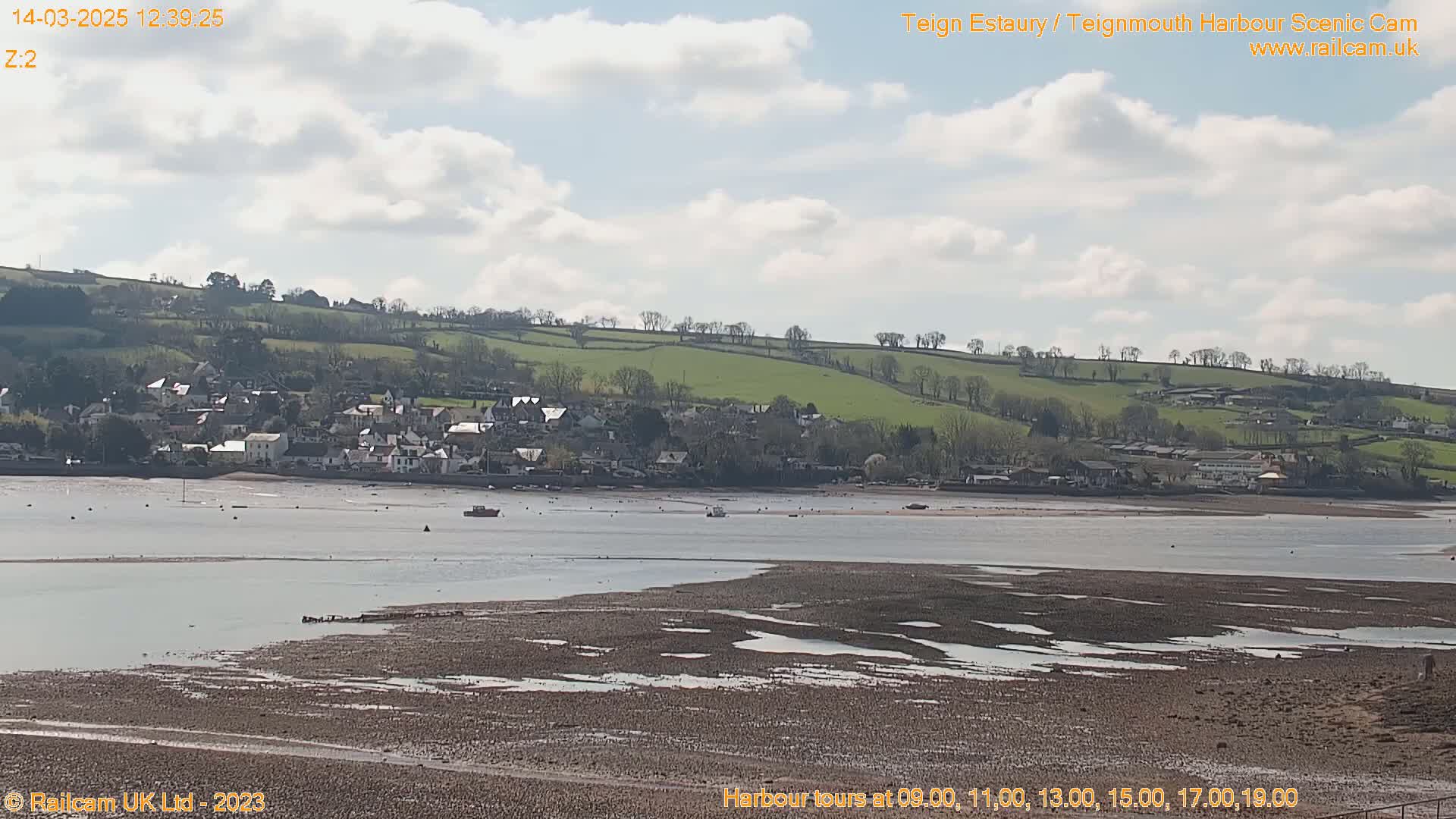 A partly cloudy day reveals a view of a town nestled beside a broad estuary with exposed mudflats at low tide, backed by rolling green hills.