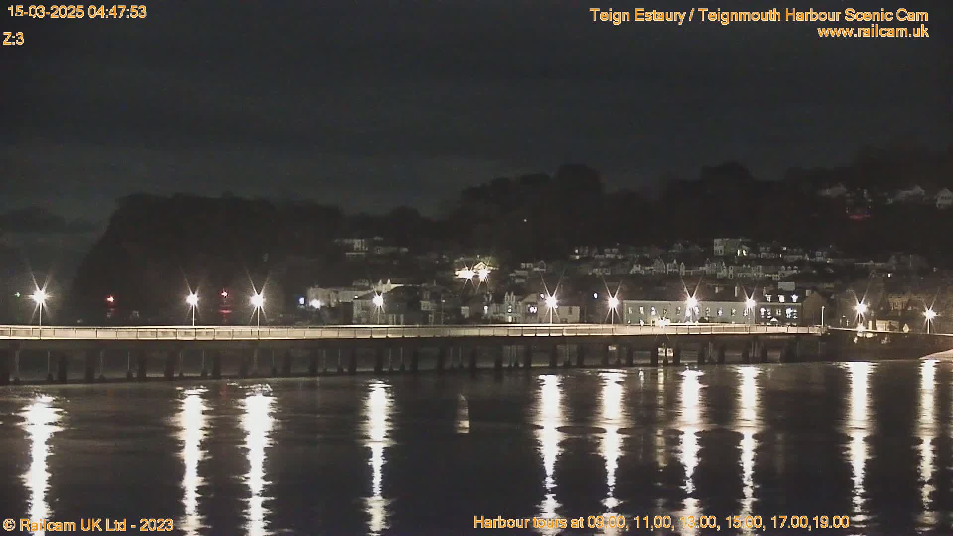 A bridge lit by streetlights spans a dark, calm body of water at night, with a town visible on the far shore.