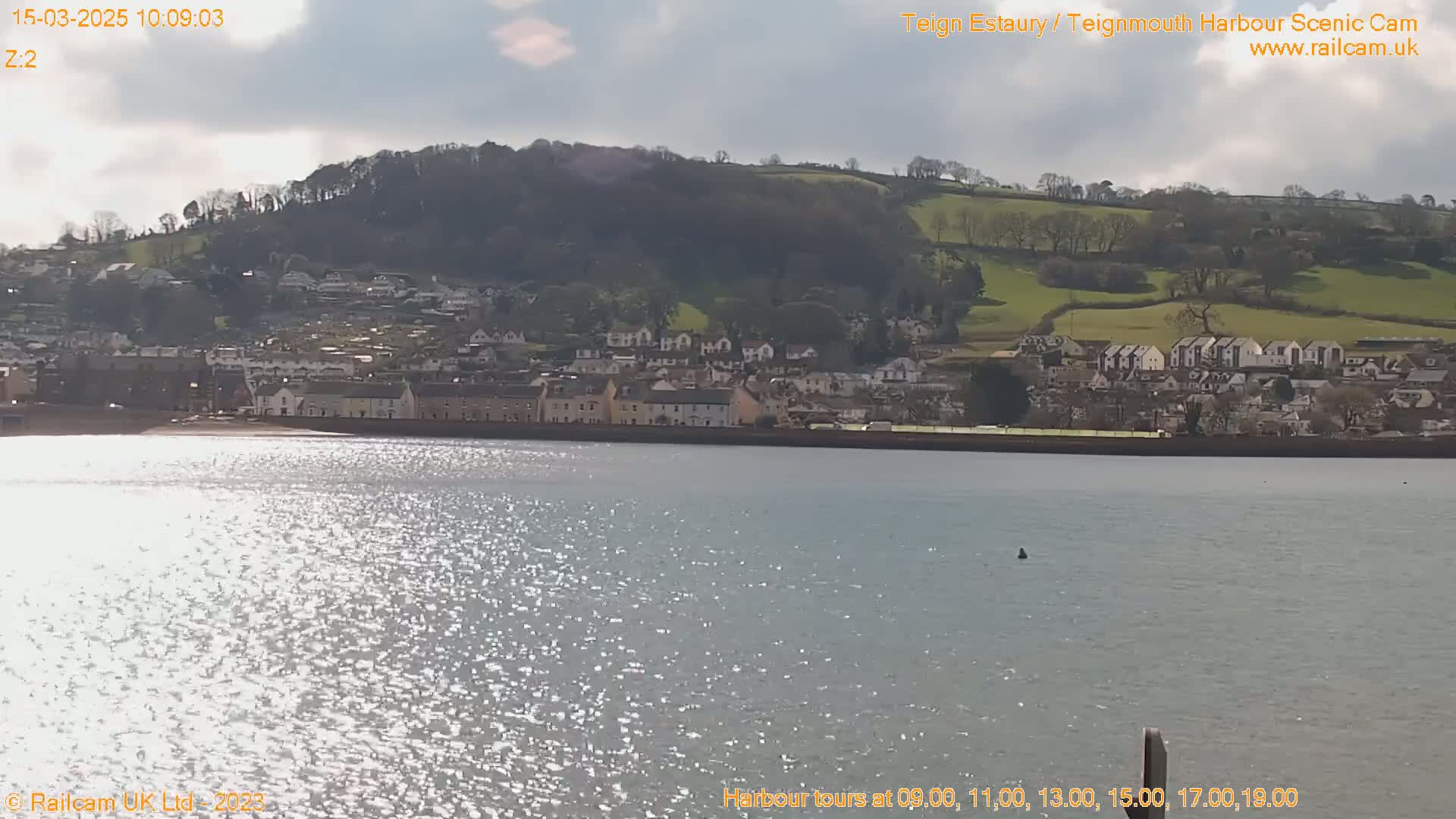 A sparkling body of water reflects sunlight, showcasing a town nestled against a hillside under a partly cloudy sky.