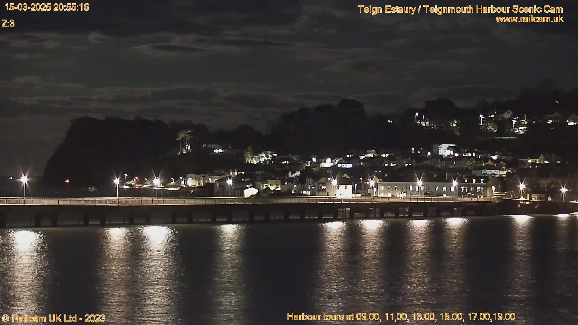 A nighttime view of a town across a calm body of water, illuminated by streetlights on a pier, under a partly cloudy sky.