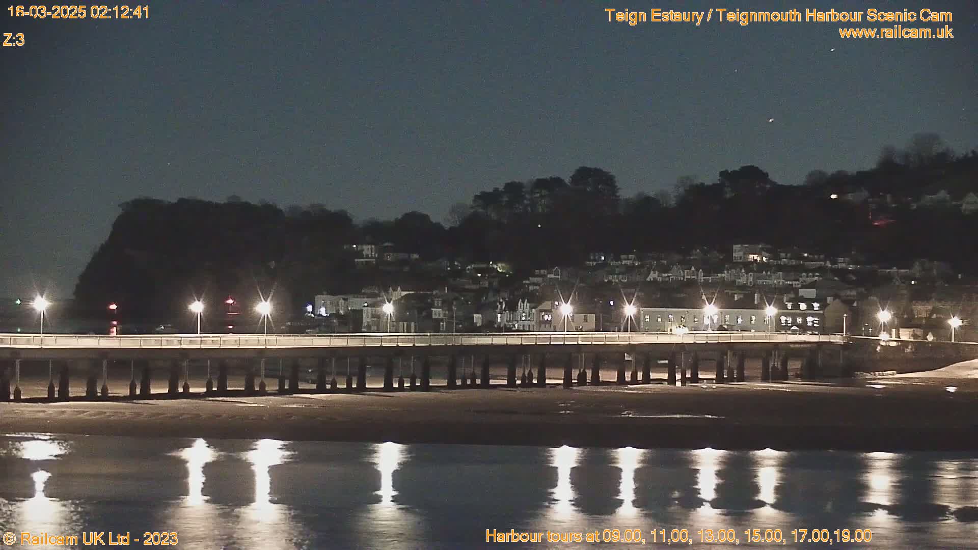 A long pier with lights extends over a calm, dark body of water reflecting the lights at night, with a hillside town visible in the background under a clear, dark sky.