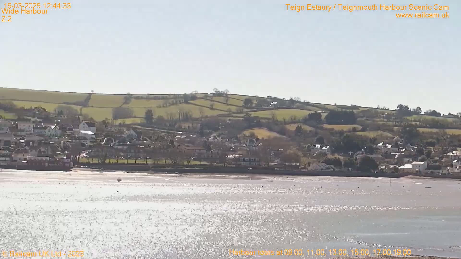 A sparkling body of water reflects sunlight, stretching out before a small town nestled at the foot of rolling, green hills under a clear, sunny sky.