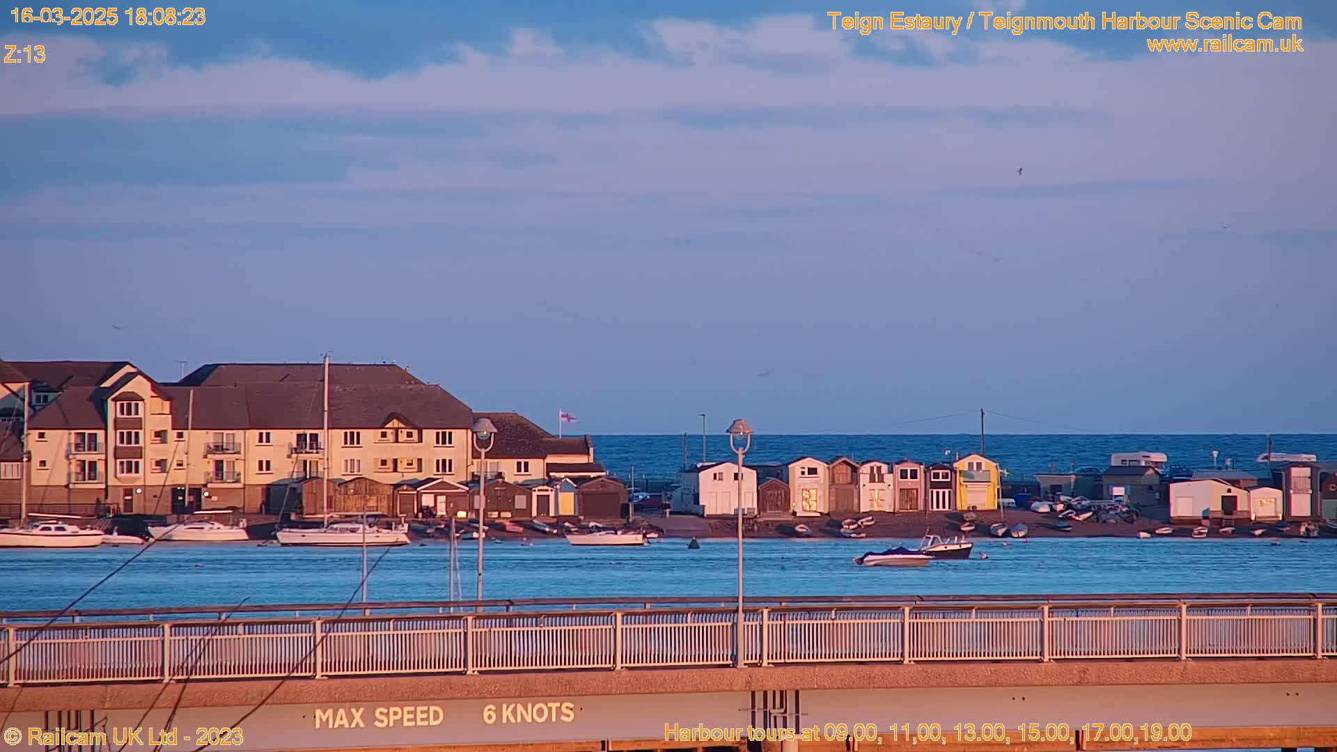 A calm, twilight scene shows a harbor with boats, buildings, and beach huts, viewed from a bridge overlooking the water.