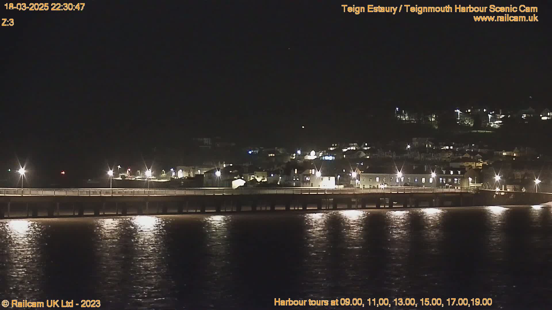 A long pier stretches across dark water reflecting lights from a town on a hill at night.