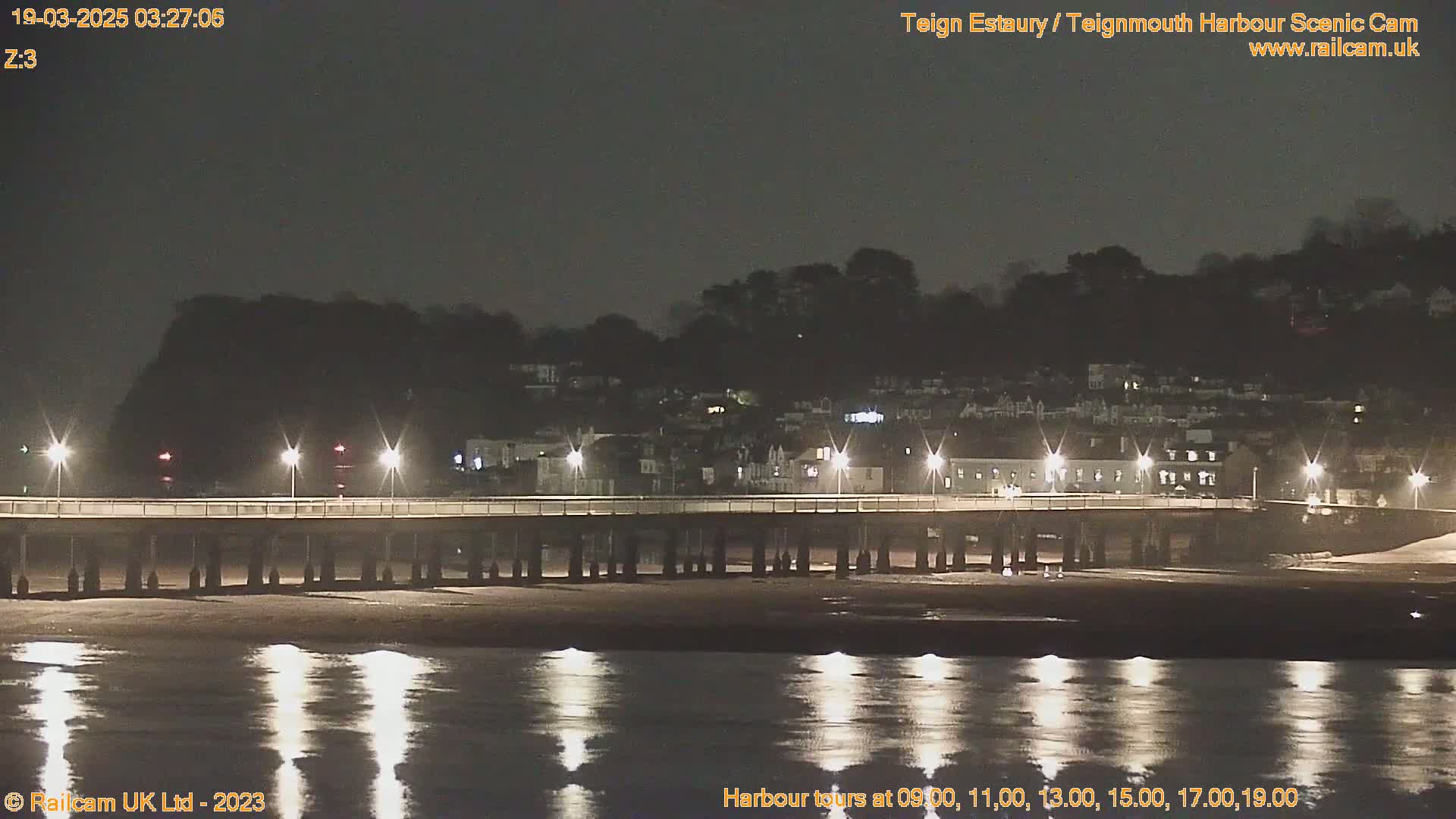 A long pier stretches across a calm, dark body of water at night, with lights illuminating a town nestled on a hillside in the background.