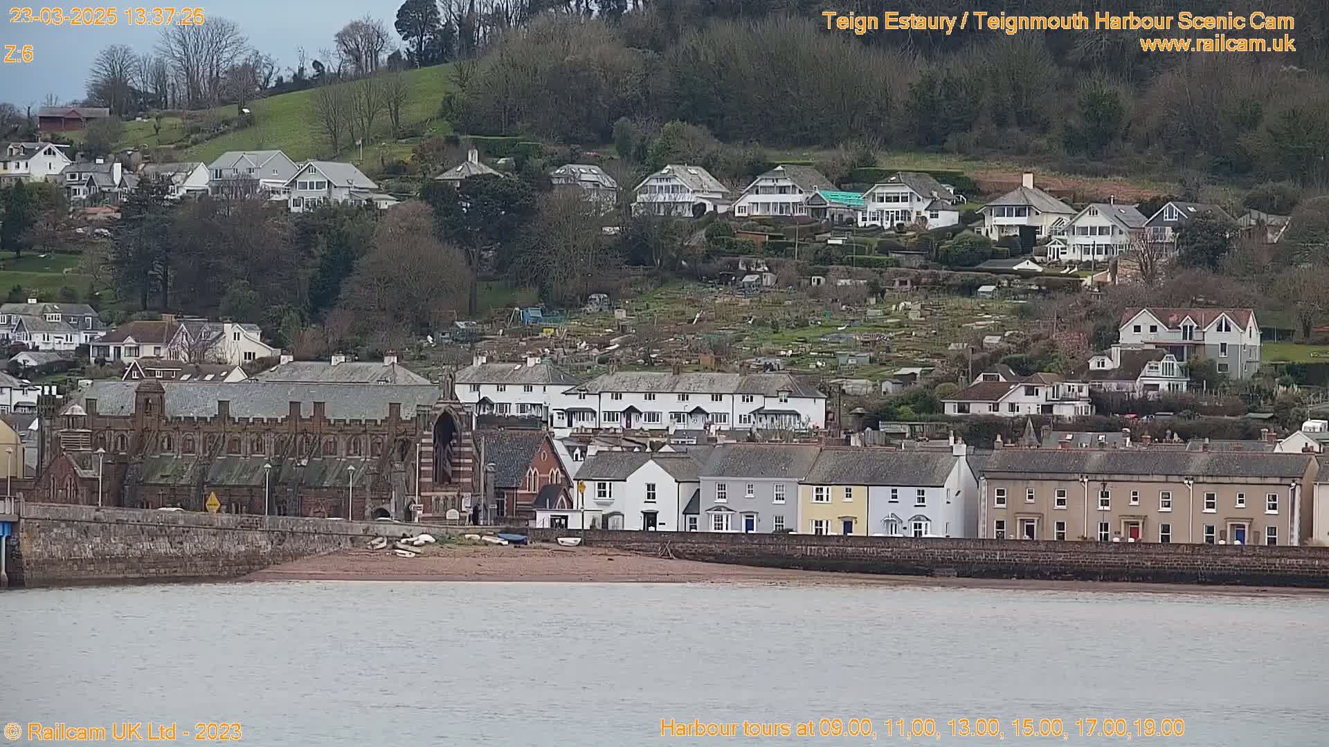 A coastal town with many houses and a large brick building sits on a hillside overlooking a calm body of water under a cloudy sky.