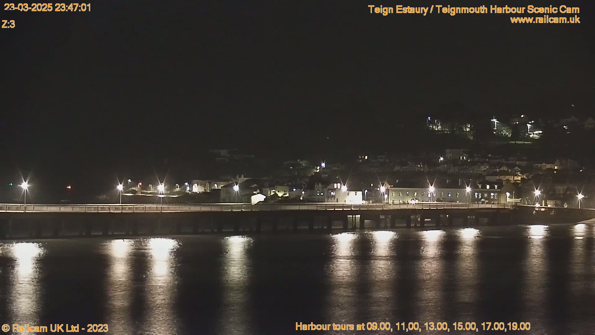 A long pier is illuminated at night, extending over calm, dark water reflecting the lights of a town on a hillside in the background.