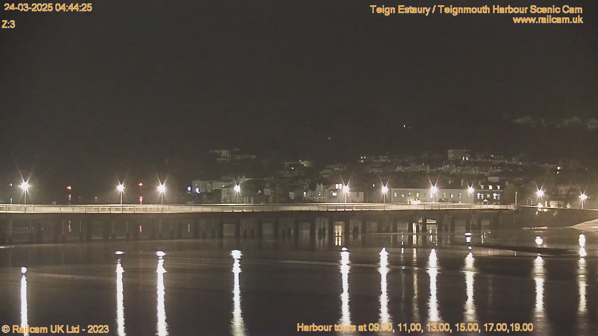 A long bridge with lights is illuminated at night, reflecting in calm water under a dark sky.