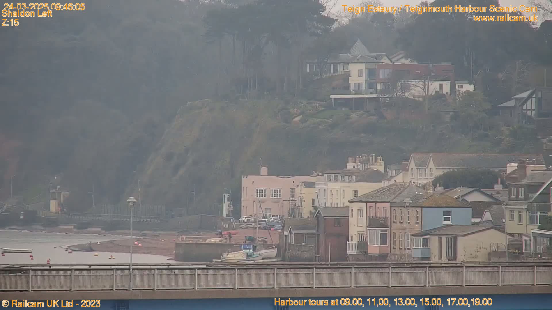A hazy view of a harbor town nestled against a hillside, with several boats moored near a low-lying sandy beach.