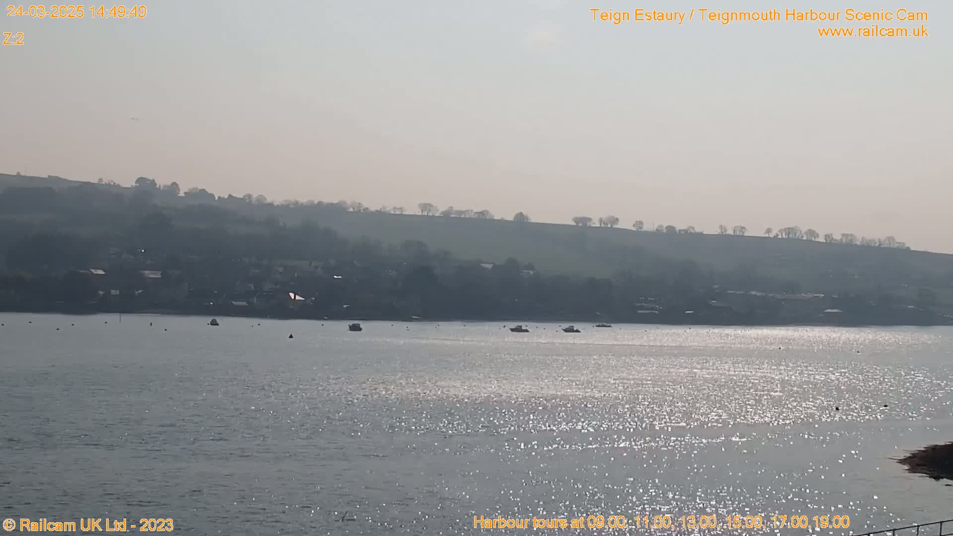 A hazy, sunny day reveals a calm body of water with several small boats, a distant shoreline with buildings and hills.