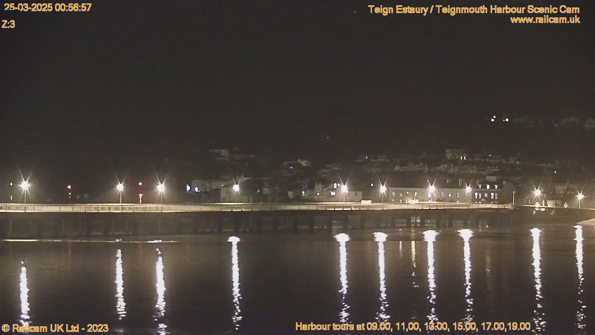 At night, a bridge with lights is reflected in calm water, with a town visible in the background.