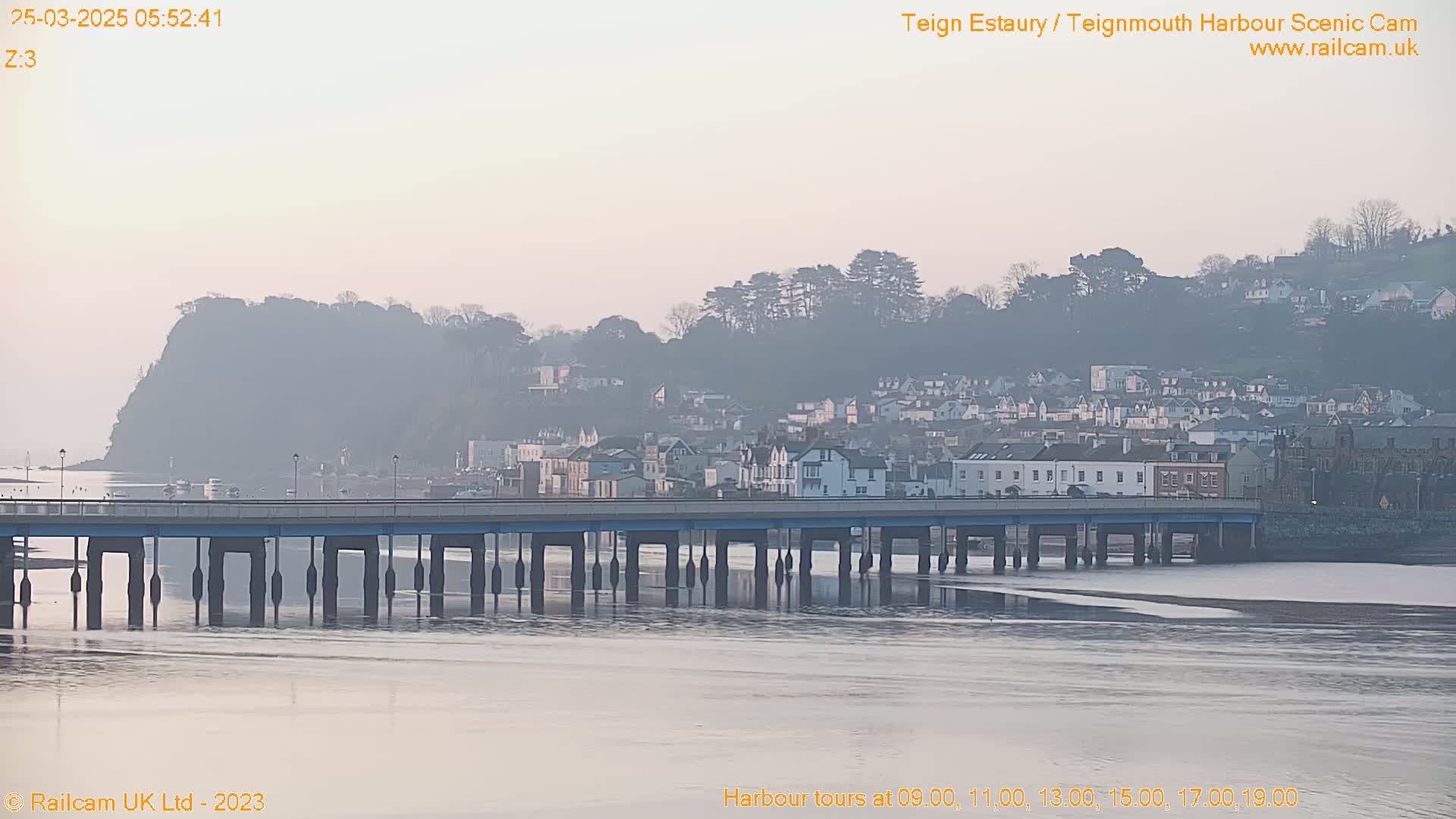 A hazy sunrise illuminates a town nestled on a hillside overlooking a calm body of water spanned by a bridge.
