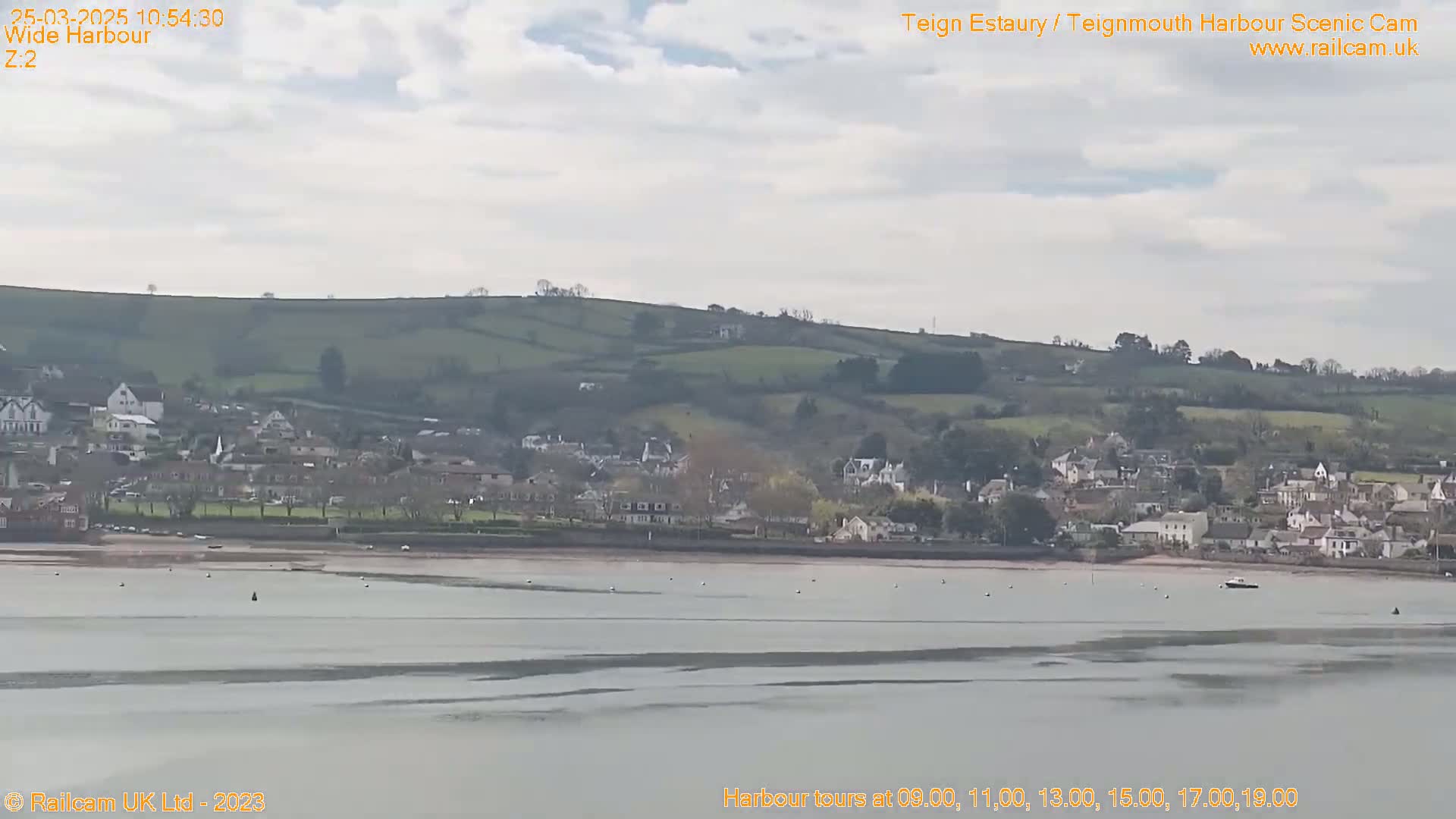 A wide shot shows a coastal town nestled against rolling green hills under a mostly cloudy sky, with a calm, wide expanse of water in the foreground.