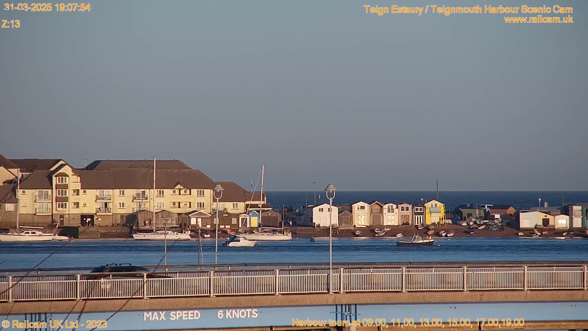 A calm, twilight-lit harbor scene shows a row of colorful beach huts, boats in the water, and a multi-story building in the background.