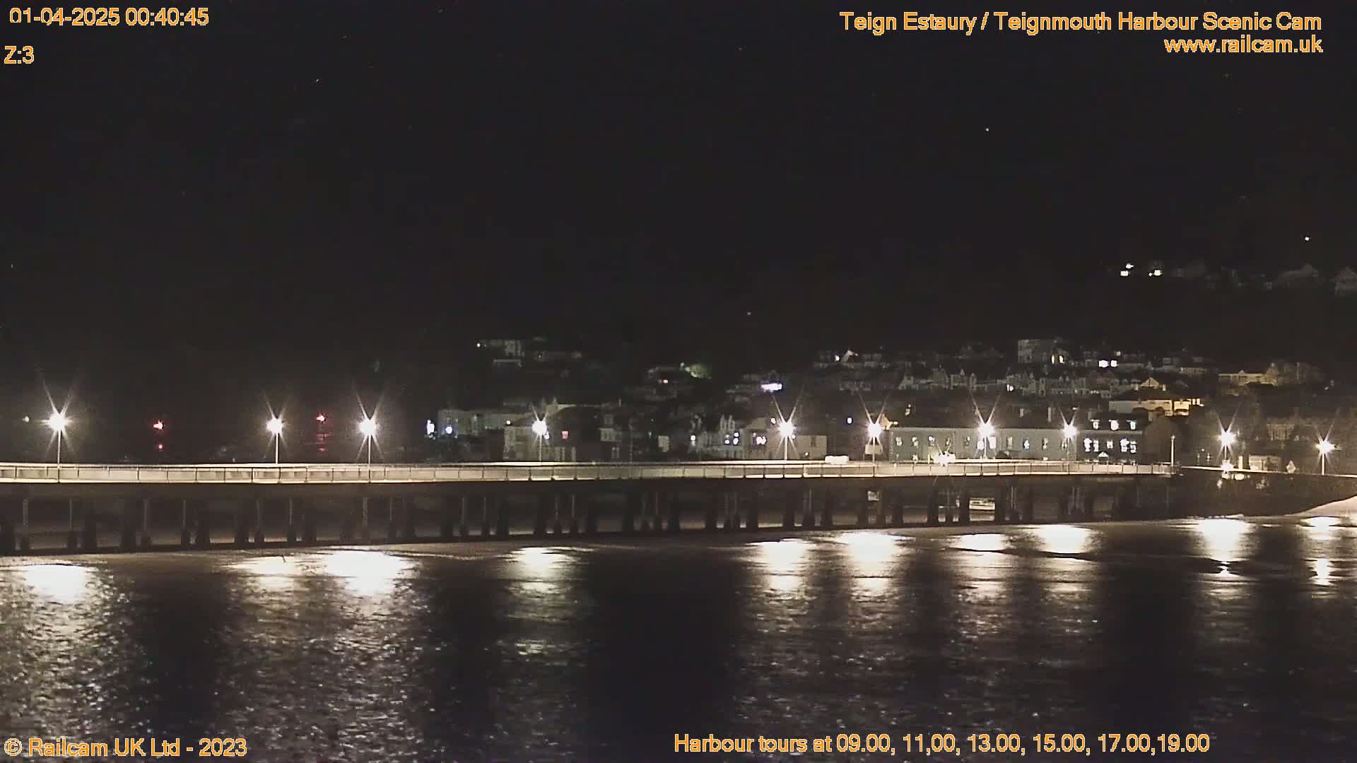 A long pier is illuminated at night, extending over dark water reflecting the lights, with a town visible in the background under a clear, dark sky.