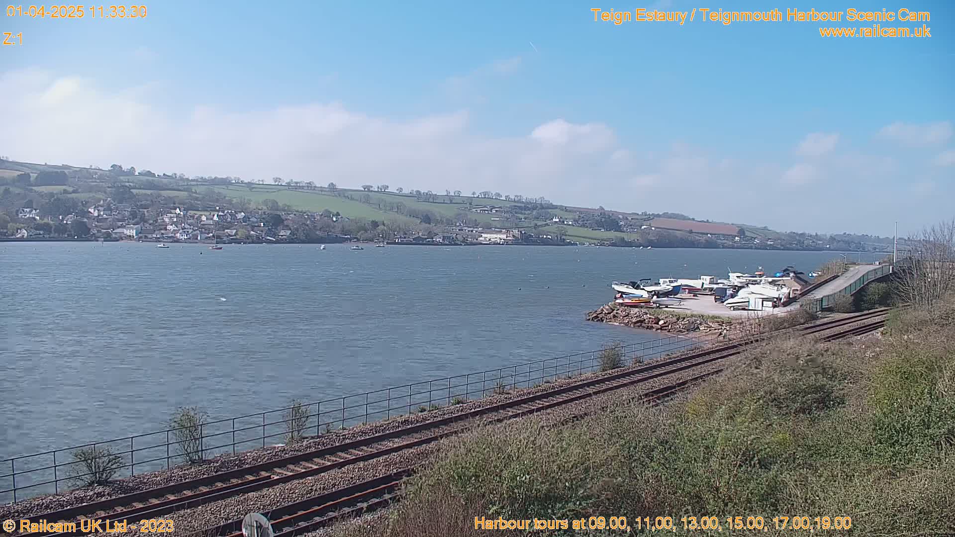 A body of water with several boats docked near a railway line under a mostly clear blue sky.