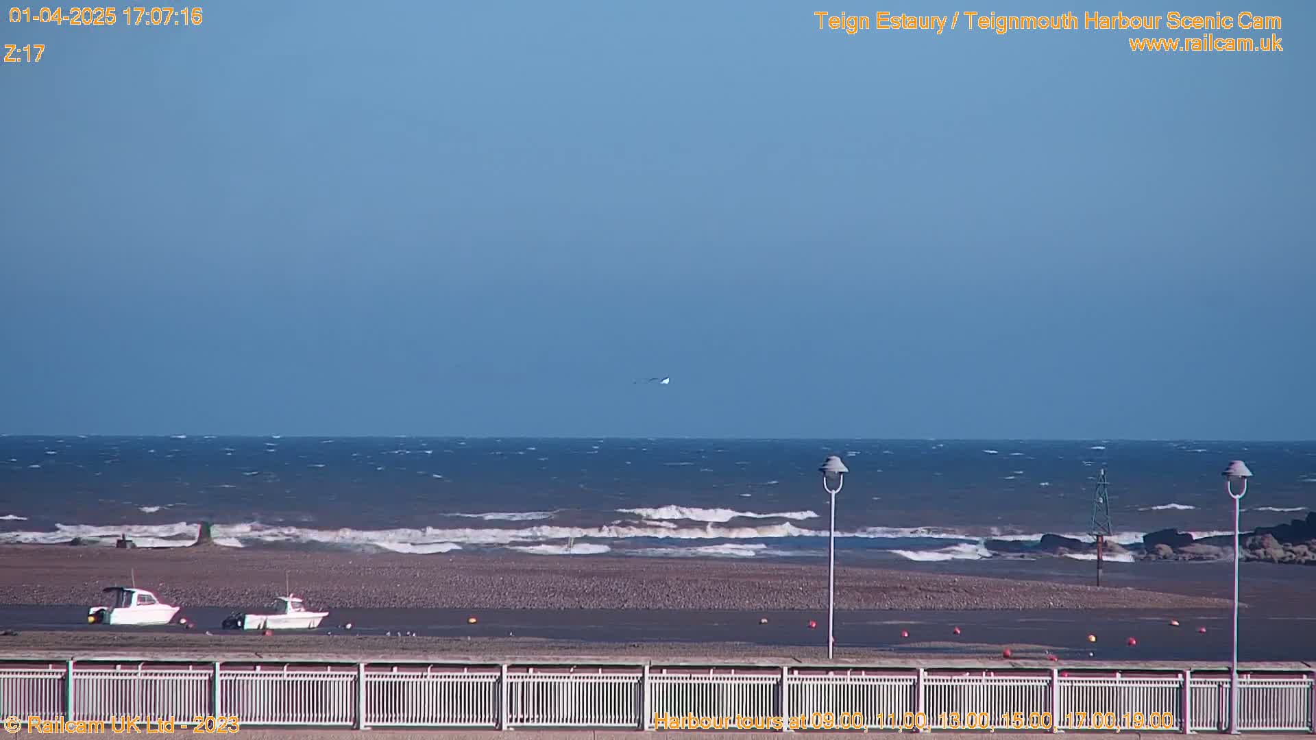 A calm, sunny day at the coast shows a wavy ocean meeting a sandy beach, two small boats are in the shallows, and a fence separates the viewer from the scene.