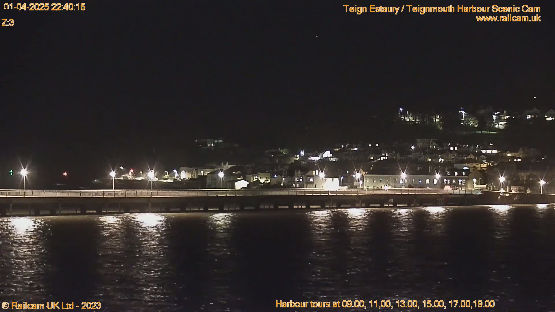 A nighttime view of a town's waterfront, illuminated by lights, across a dark body of water from a pier.