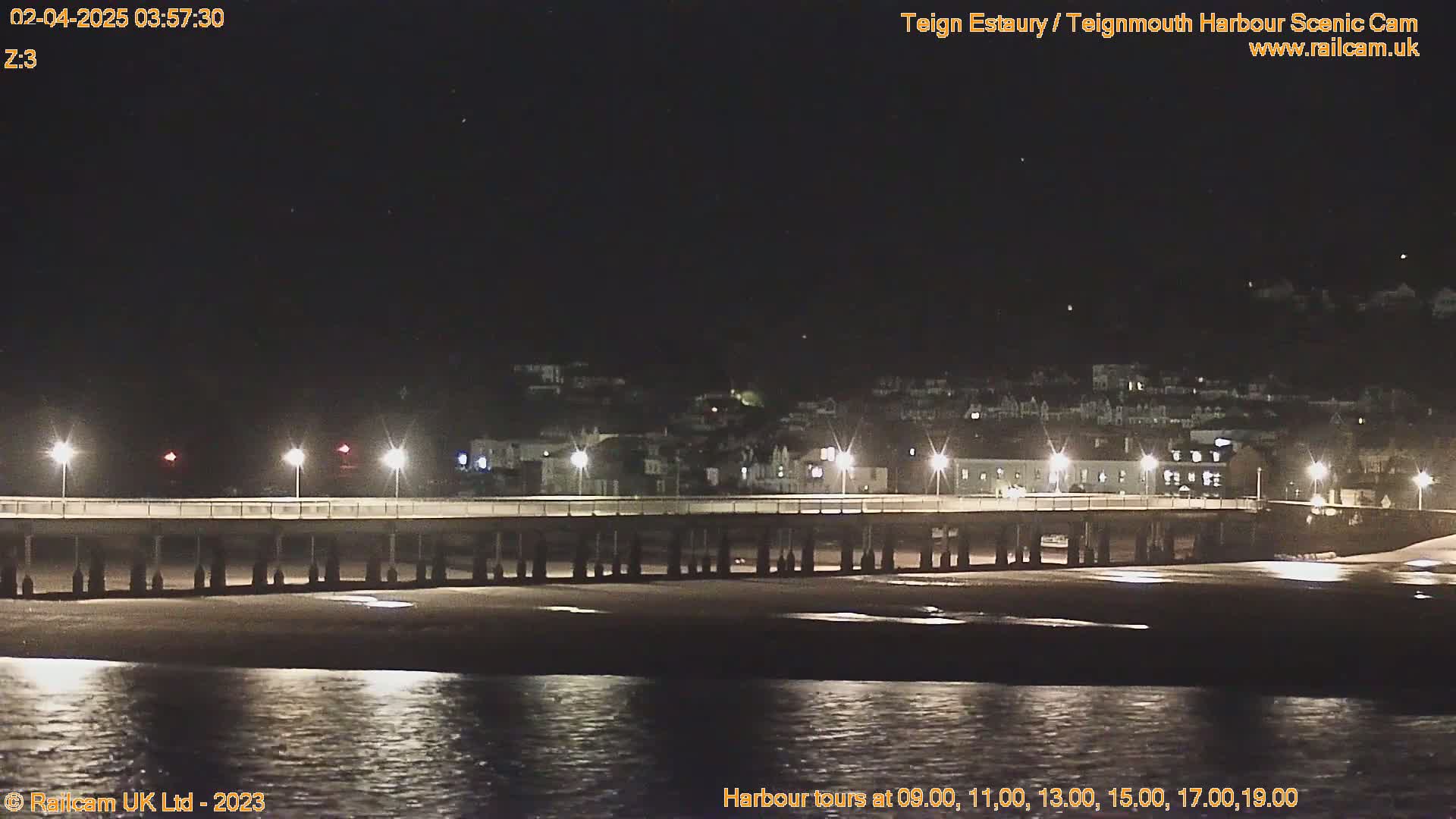 A long pier with lights extends over a dark, calm body of water at night, with a town illuminated on the far shore.