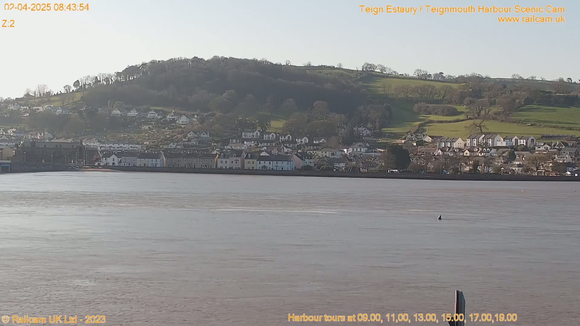 A calm body of water stretches out before a town nestled against a gently sloping hillside under a clear, bright sky.