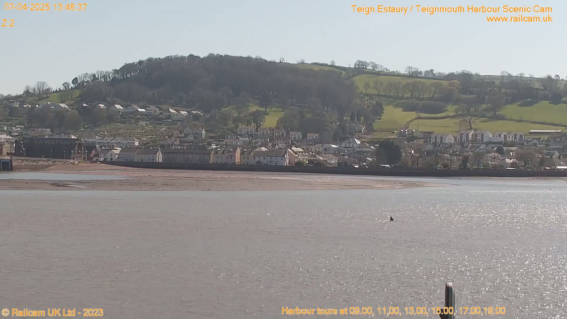 A coastal town sits on a hillside overlooking a calm, sunny estuary.
