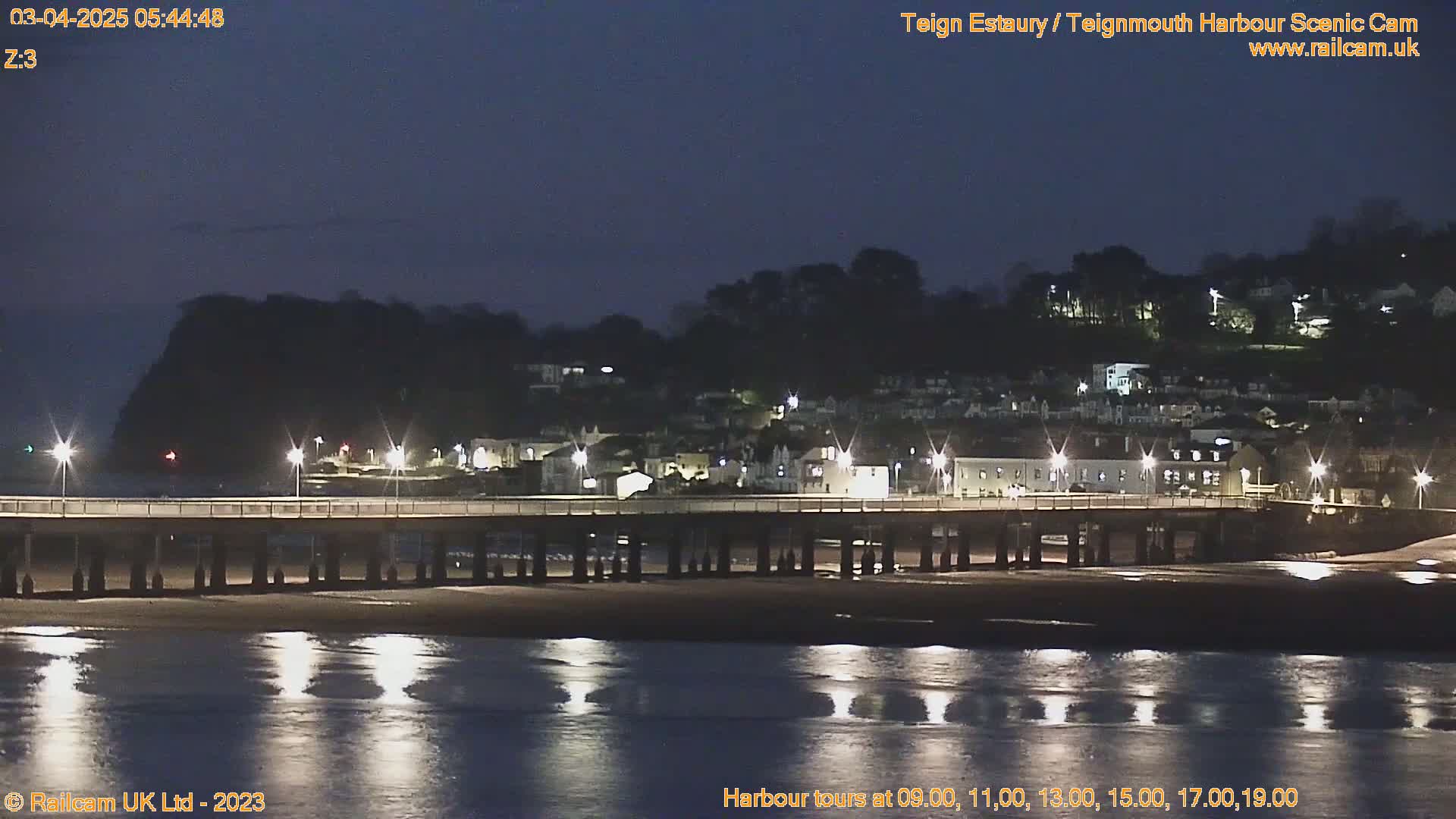 A long pier stretches across a calm, dark river reflecting the lights of a hillside town at night.
