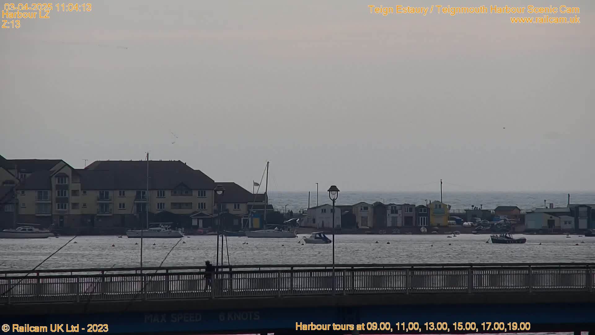 A view from a bridge shows a calm harbor with several boats, buildings, and a person walking along the bridge under an overcast sky.