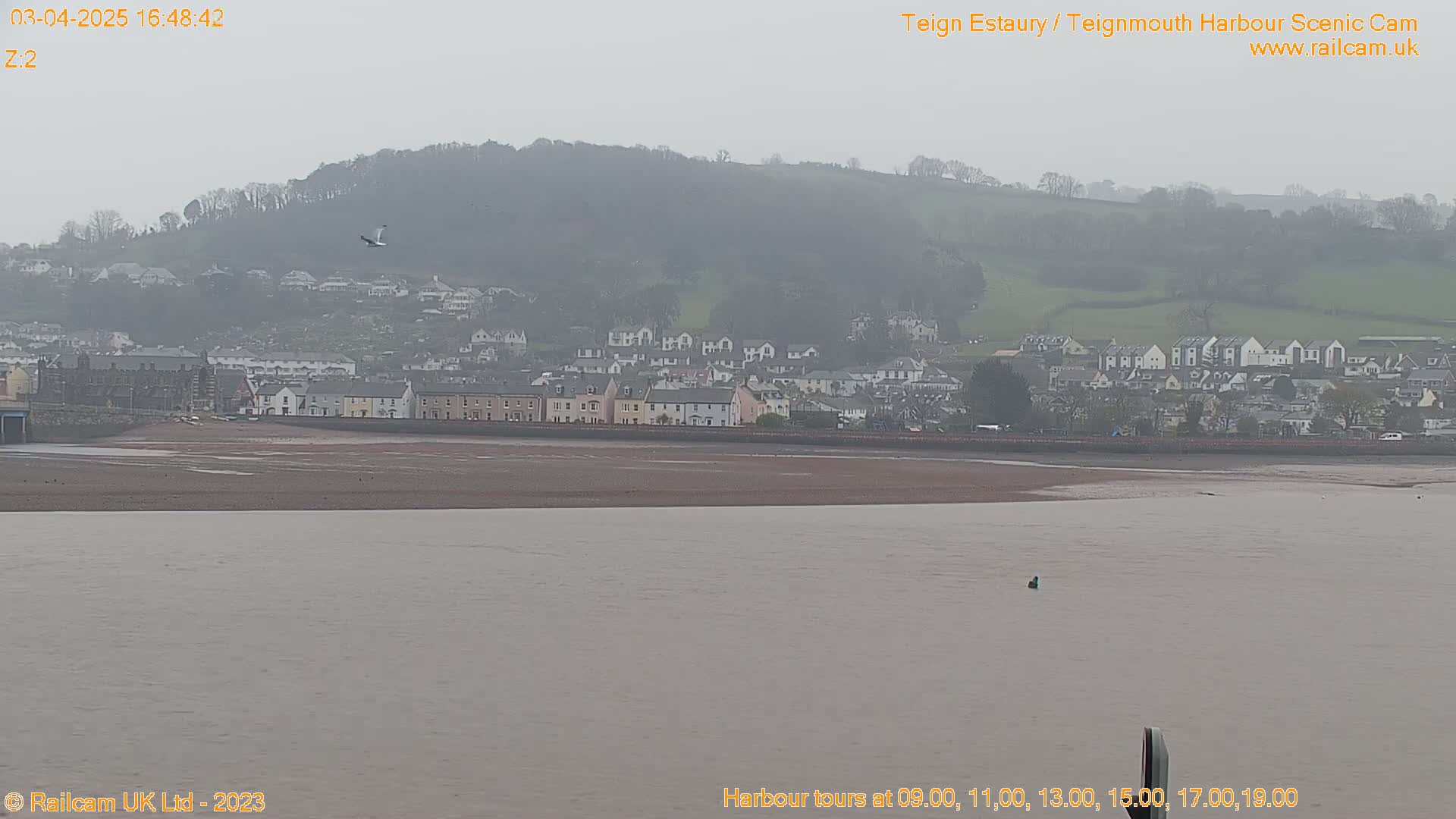 A hazy view shows a coastal town nestled against a hillside, with a wide, calm body of water in the foreground.