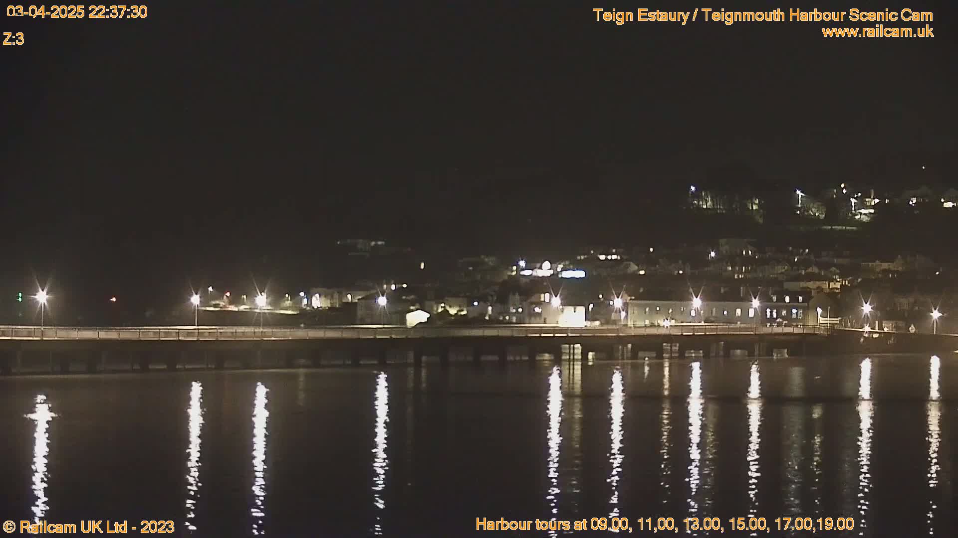 A nighttime view of a calm body of water reflecting the lights of a town across a bridge under a clear, dark sky.