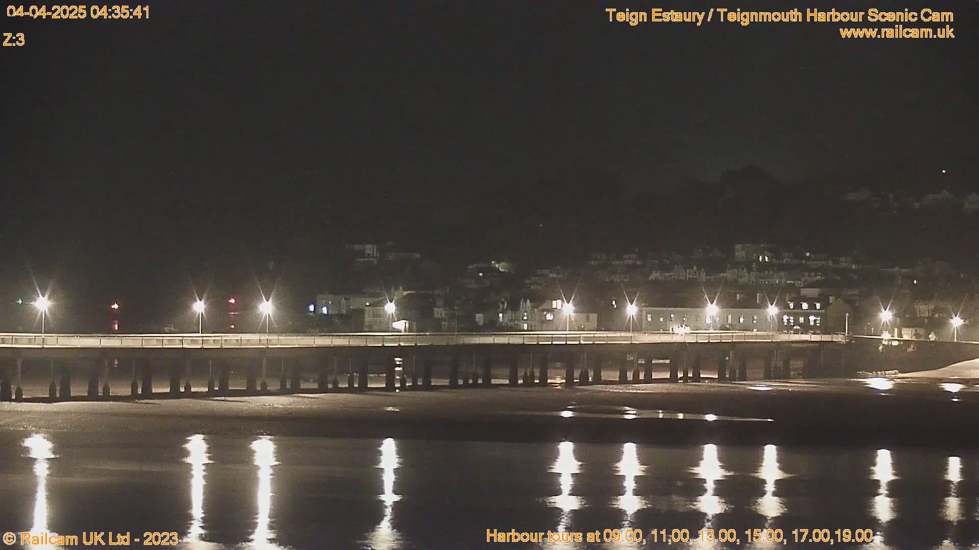 A long pier with lights illuminates a dark, calm body of water at night, with a town visible in the background.
