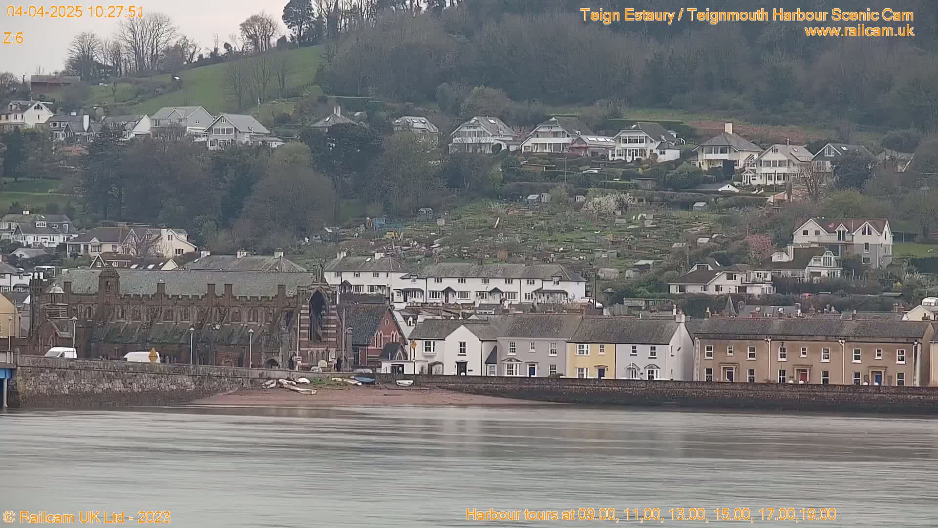 A coastal town with numerous houses and buildings sits on a hillside overlooking a calm body of water under an overcast sky.