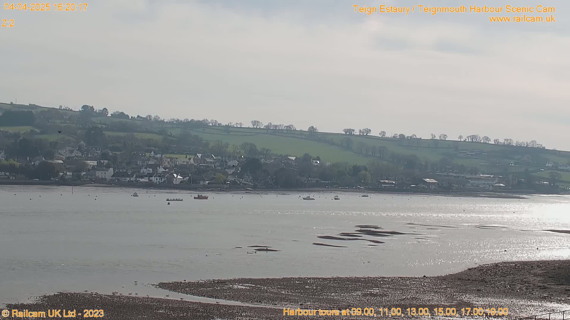 A calm, bright day shows a wide estuary with a town in the background, several small boats on the water, and a muddy shoreline in the foreground.
