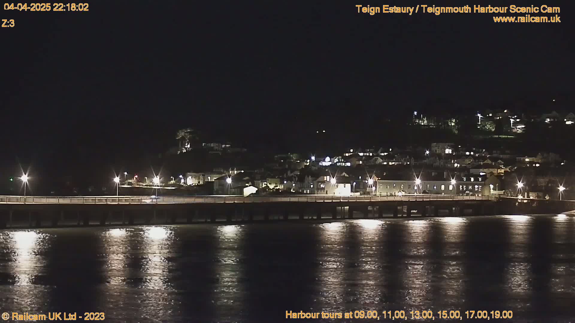 A long pier stretches across dark water at night, illuminated by lights reflecting on the calm surface, with a hillside town visible in the background.