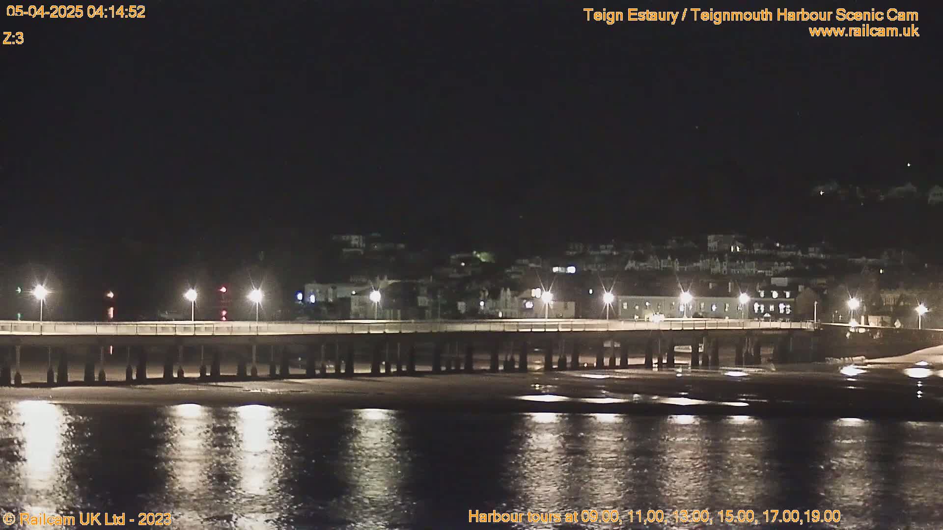 A long pier is illuminated at night, extending over dark water reflecting the lights, with a town visible in the background under a clear, dark sky.